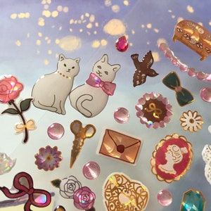 May include: A sheet of colorful stickers featuring cats, flowers, bows, and other decorative elements. The stickers are on a light blue background with a glittery texture.
