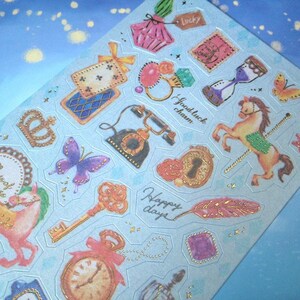 May include: A sheet of colorful stickers with a blue background. The stickers feature various designs, including a butterfly, a crown, a key, a clock, a feather, a horse, and a telephone. The stickers have gold accents and the text "Collect happiness", "Good luck charm", "Happy days", and "Lucky" are visible.