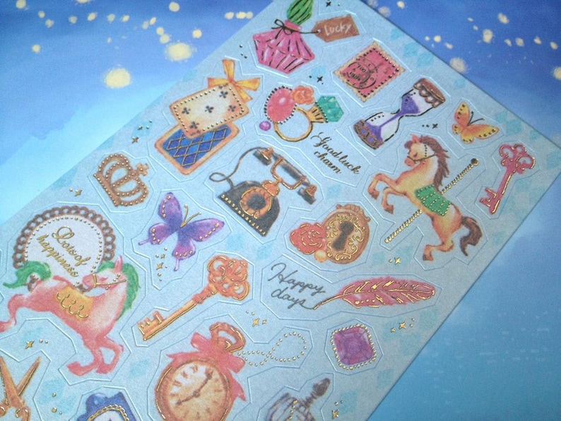 May include: A sheet of colorful stickers with a blue background. The stickers feature various designs, including a butterfly, a crown, a key, a clock, a feather, a horse, and a telephone. The stickers have gold accents and the text "Collect happiness", "Good luck charm", "Happy days", and "Lucky" are visible.