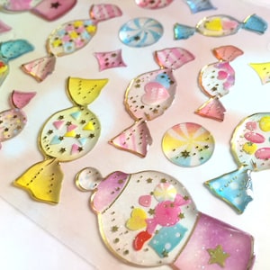 May include: A sheet of colorful, transparent, candy-shaped stickers with gold glitter accents. The stickers are shaped like lollipops, candy canes, and wrapped candies.