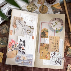 May include: An open notebook with decorative paper stickers featuring Japanese-inspired designs. The stickers include images of flowers, birds, landscapes, and calligraphy. The notebook is open to a page with a sticker that reads "保持熟愛".