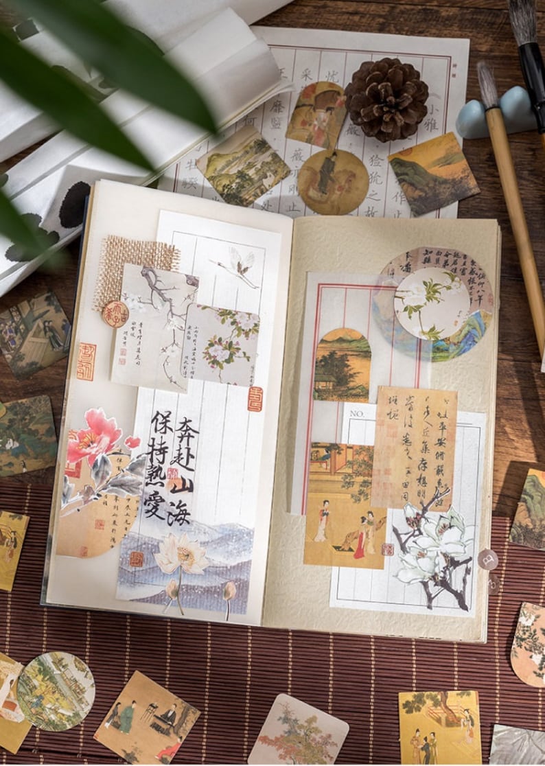 May include: An open notebook with decorative paper stickers featuring Japanese-inspired designs. The stickers include images of flowers, birds, landscapes, and calligraphy. The notebook is open to a page with a sticker that reads "保持熟愛".