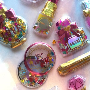 May include: Assortment of colourful, glittery, and holographic resin stickers with various makeup-themed designs. The stickers feature words like "Love", "Sweet", and "Eye Shadow".
