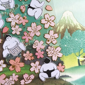 May include: A sheet of colourful stickers featuring illustrations of sumo wrestlers and pink cherry blossoms. The background is a green and blue watercolour painting of a mountain and a tree.