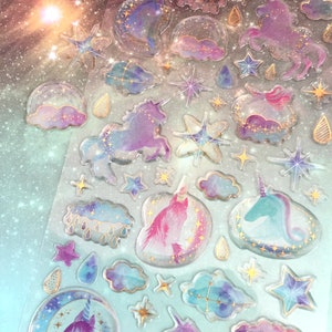 May include: A sheet of clear, iridescent stickers featuring a variety of unicorn designs. The stickers are decorated with gold glitter, stars, and clouds. The unicorns are in various poses, including running, standing, and looking up.