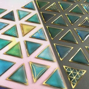 Puede incluir: Primer plano de pegatinas triangulares decorativas con borde dorado. Los triángulos presentan tonos de verde azulado, azul y verde con patrones geométricos dorados. Las pegatinas están dispuestas sobre un fondo blanco y gris.
