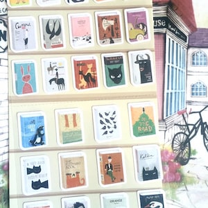 Puede incluir: Una hoja de pegatinas de temática de libros con ilustraciones coloridas de varios animales, objetos y texto. Las pegatinas presentan títulos como "El Gato Negro", "Primeros Auxilios", "El Libro del Elefante" y "En el Camino".