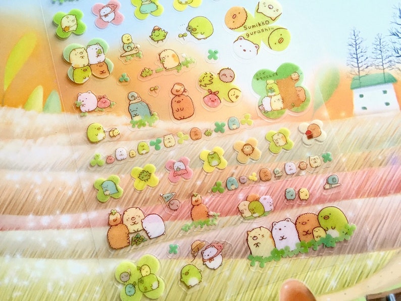 May include: A sheet of colorful, transparent stickers featuring adorable cartoon characters. The stickers are arranged in rows and include various animals, plants, and objects. The text "Sumikko Gurashi" is visible at the top of the sheet.