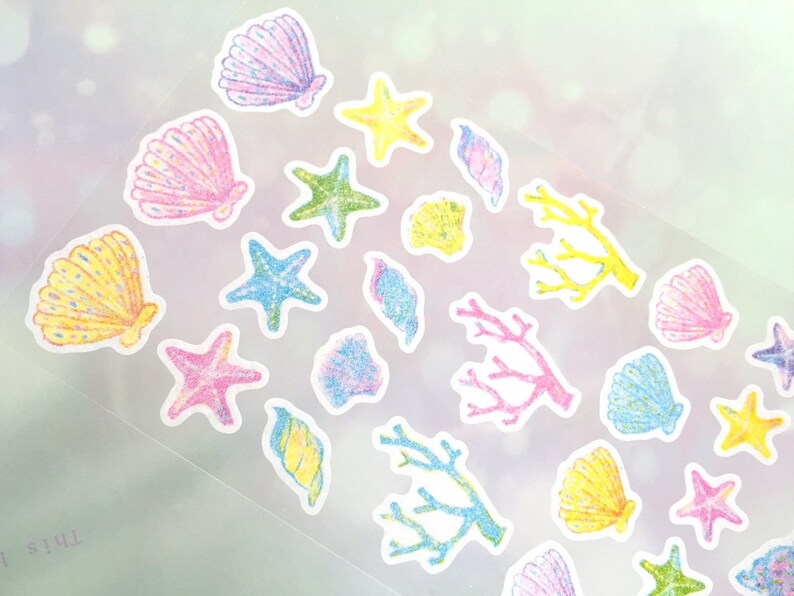 May include: A sheet of colorful stickers featuring various sea-themed designs, including seashells, starfish, and coral. The stickers are outlined in white and have a glittery texture.