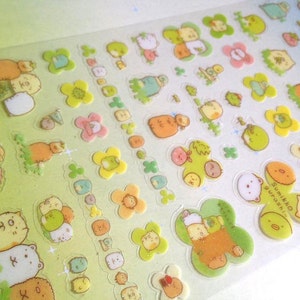 May include: A sheet of colorful stickers featuring cartoon characters. The stickers are on a white background with green, yellow, and pink accents. The characters are all different shapes and sizes, and some have text on them. The text on the stickers says "Sumikko Gurashi".