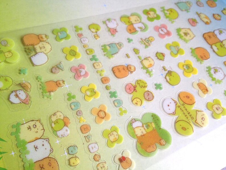 May include: A sheet of colorful stickers featuring cartoon characters. The stickers are on a white background with green, yellow, and pink accents. The characters are all different shapes and sizes, and some have text on them. The text on the stickers says "Sumikko Gurashi".