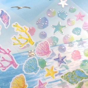 May include: A sheet of colorful stickers featuring seashells, starfish, and coral. The stickers are on a clear background with a light blue and white watercolor design.