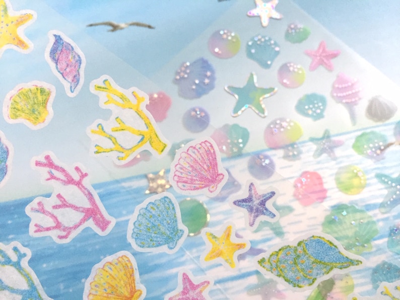 May include: A sheet of colorful stickers featuring seashells, starfish, and coral. The stickers are on a clear background with a light blue and white watercolor design.