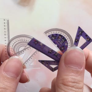 Peut inclure: Un ensemble de quatre rapporteurs en plastique violet avec des personnages de dessins animés imprimés dessus. Les rapporteurs sont en forme de demi-cercle, de triangle et de deux carrés.