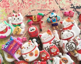 May include: A collection of colorful, 3D, lucky cat figurines with Japanese characters. The figurines are made of a soft, rubbery material and have a variety of designs, including a red and white cat with a gold coin, a white cat with a red and white scarf, and a black cat with a red and white scarf.
