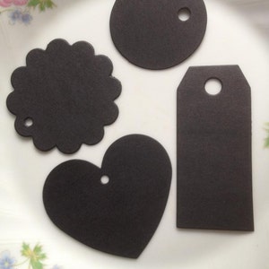 May include: Four black paper gift tags in different shapes: a circle, a heart, a rectangle, and a scalloped circle. Each tag has a hole punched in the top for string or ribbon.