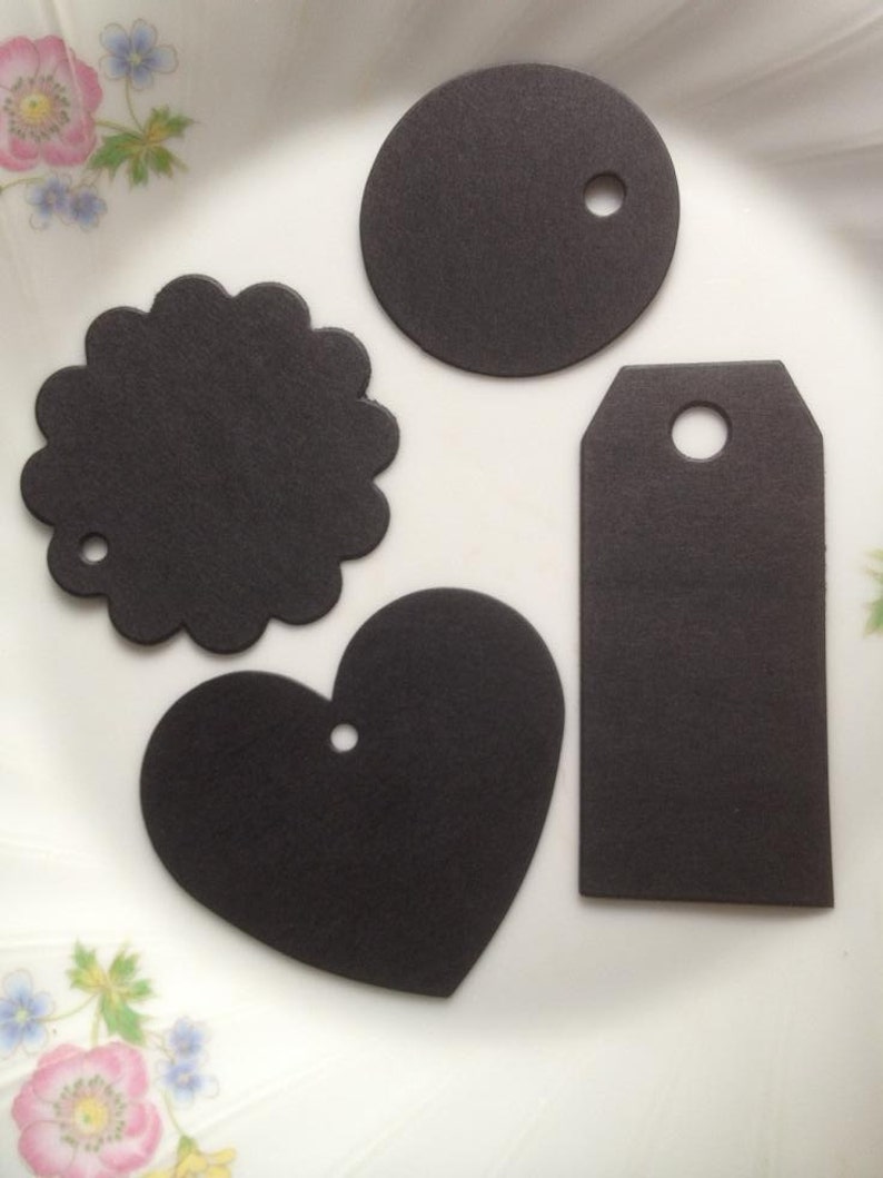 May include: Four black paper gift tags in different shapes: a circle, a heart, a rectangle, and a scalloped circle. Each tag has a hole punched in the top for string or ribbon.