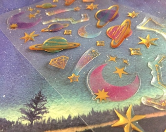 May include: A sheet of clear stickers with a space theme. The stickers feature gold foil accents and include planets, stars, moons, and a comet.