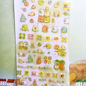 May include: A sheet of clear stickers featuring various adorable cartoon characters from the Sumikko Gurashi series. The characters are mostly white and brown with some green and yellow accents. The stickers are arranged in rows and columns on a white background. The text "Sumikko Gurashi" is printed in black on a white sticker in the center of the sheet.