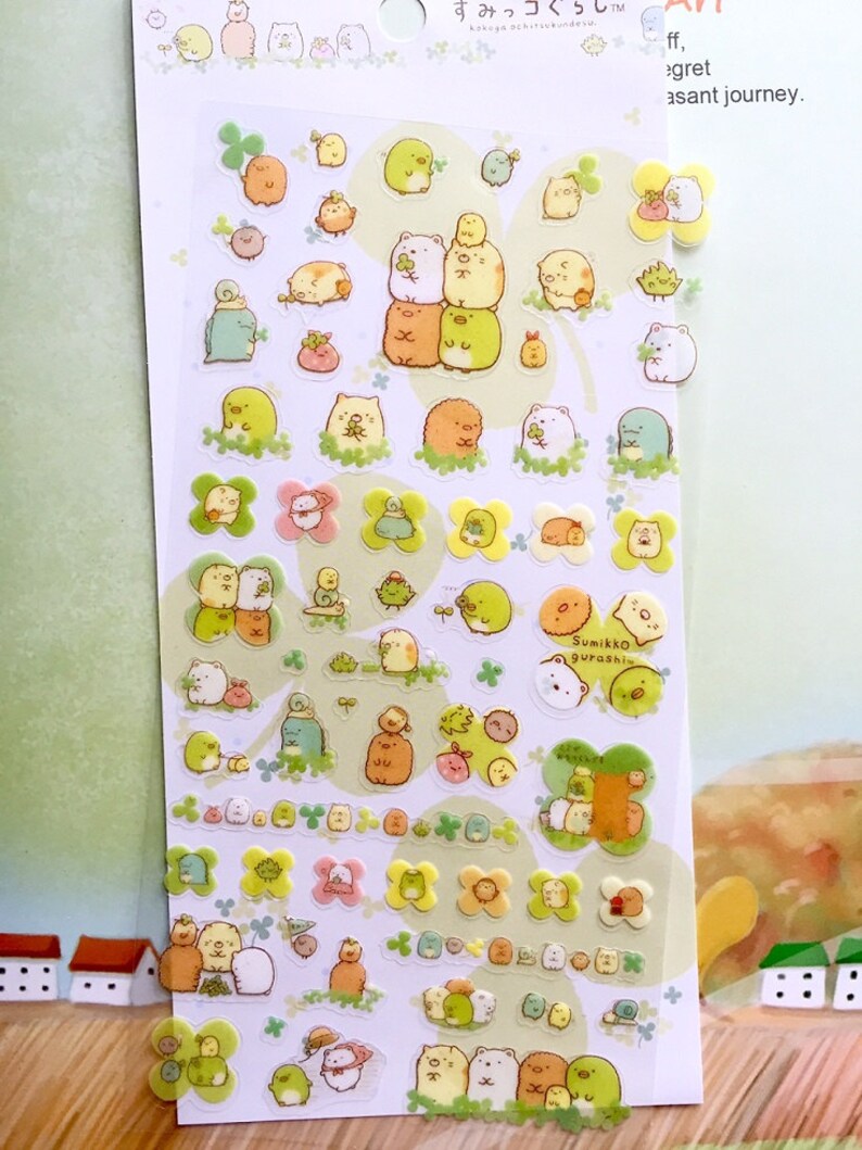 May include: A sheet of clear stickers featuring various adorable cartoon characters from the Sumikko Gurashi series. The characters are mostly white and brown with some green and yellow accents. The stickers are arranged in rows and columns on a white background. The text "Sumikko Gurashi" is printed in black on a white sticker in the center of the sheet.