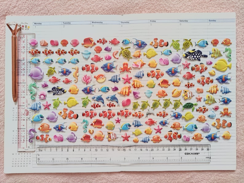 Puede incluir: Una hoja de pegatinas brillantes y coloridas con varias criaturas marinas, incluyendo peces, caballitos de mar, estrellas de mar y tortugas. Las pegatinas est&aacute;n dispuestas en filas sobre un fondo blanco con una regla en la parte inferior.