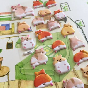 May include: A collection of 17 colorful hamster stickers with various poses and expressions. The stickers are on a white background with a painted scene of a bookshelf, a cactus, and a green couch.