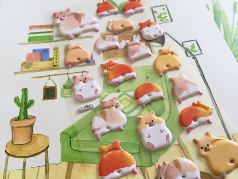 May include: A collection of 17 colorful hamster stickers with various poses and expressions. The stickers are on a white background with a painted scene of a bookshelf, a cactus, and a green couch.
