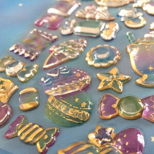 Puede incluir: Una hoja de pegatinas holográficas iridiscentes con varias formas y diseños, incluyendo estrellas, caramelos y una luna creciente. Las pegatinas tienen un contorno dorado y son perfectas para agregar un toque de brillo a las manualidades, los diarios y más.