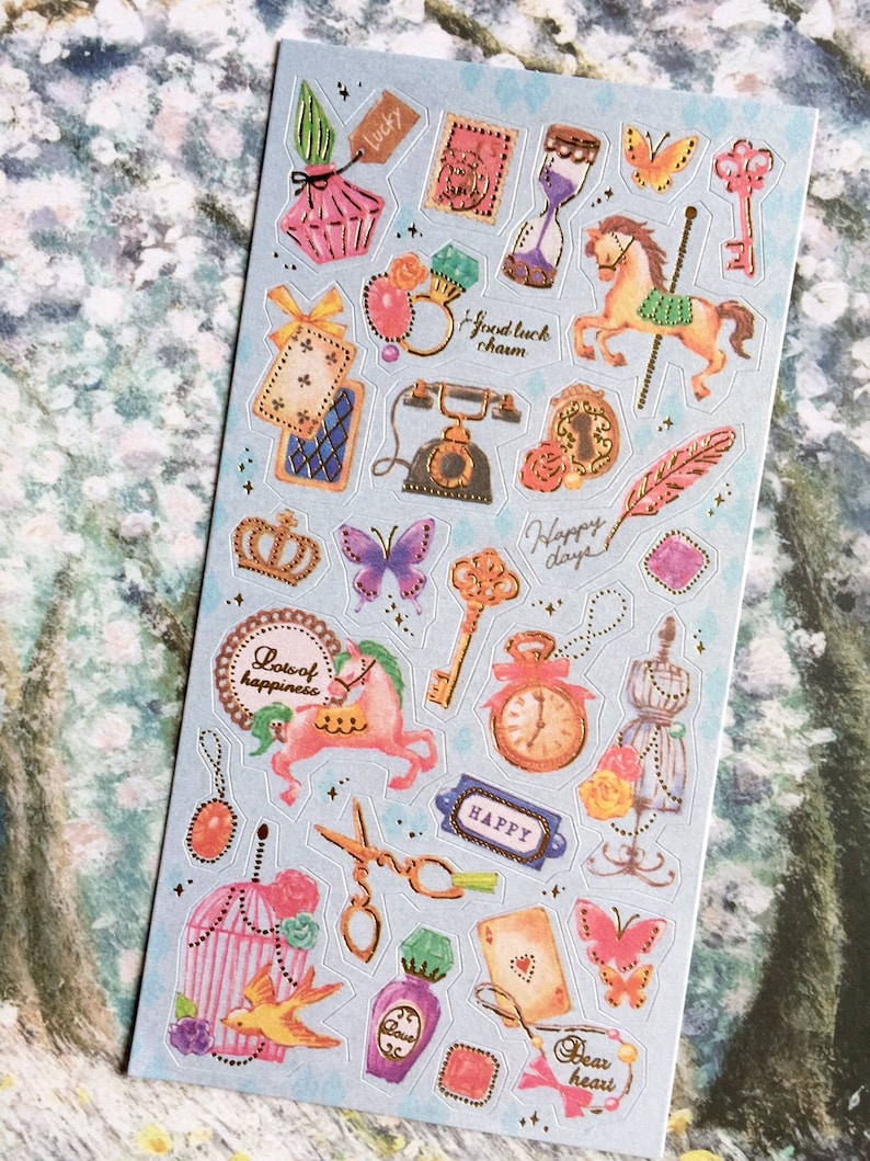 May include: A sheet of colorful stickers with a variety of designs, including butterflies, a carousel horse, a birdcage, a crown, a key, a clock, a bottle of perfume, a telephone, a scissors, a playing card, and a feather. The stickers have gold accents and are on a light blue background.