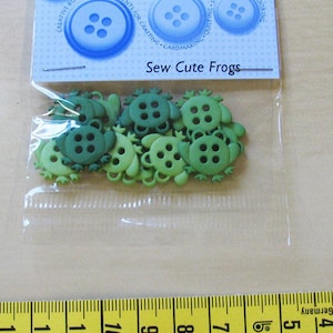 Könnte beinhalten: Eine Packung grüner, froschförmiger Knöpfe mit dem Text "Sew Cute Frogs" auf der Verpackung.
