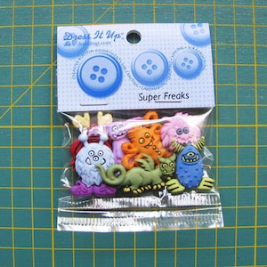 Könnte beinhalten: Eine Packung bunter Plastikknöpfe in Monsterform. Die Knöpfe befinden sich in einem durchsichtigen Plastikbeutel mit einem weißen Etikett, auf dem "Dress It Up" und "Super Freaks" steht.