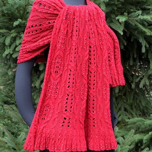 May include: A red knitted shawl with a lacy pattern and beaded accents. The shawl is draped over a mannequin and is displayed against a green background.