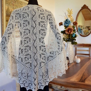 Puede incluir: Un chal de ganchillo blanco con un delicado patrón de encaje. El chal está drapeado sobre un maniquí y se muestra sobre un fondo claro.