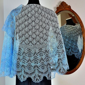 Pode incluir: Um xale de renda azul claro e branco com um padrão delicado. O xale está drapeado sobre um manequim e se reflete em um espelho.