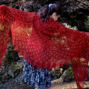 May include: A red lace shawl with a delicate pattern and a gold shimmer. The shawl is draped over a person's shoulders and is flowing in the wind.