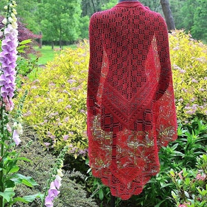 May include: A red crocheted shawl with a lacy pattern. The shawl is draped over a mannequin and is displayed against a backdrop of green foliage.