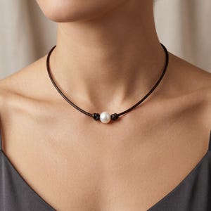 May include: A choker necklace featuring a single white spherical pearl centered on a thin dark brown leather cord. The pearl is secured in place by two small knots tied in the leather cord on either side of the pearl, creating a simple design.