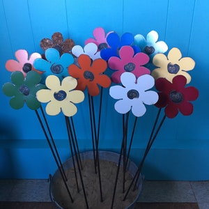 May include: A collection of colorful metal flowers in a galvanized bucket. The flowers are made of metal and have a variety of colors, including red, blue, green, yellow, and pink. The flowers are arranged in a bucket filled with sand.
