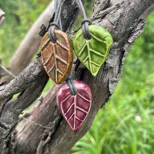 May include: Three ceramic leaf-shaped pendants in glossy orange, bright green, and deep magenta, each featuring embossed vein details. The pendants are suspended from thin grey cords, showcasing a smooth, glazed finish on each individual piece.