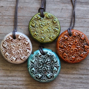 May include: Four ceramic pendant necklaces with intricate floral designs. The pendants are in shades of white, green, teal, and orange, each with a leather cord. The necklaces are displayed on a rustic wooden surface.