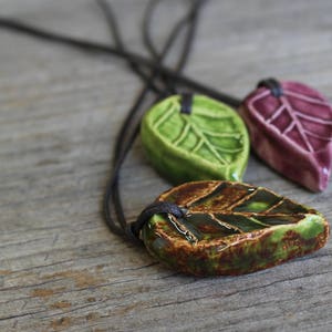 May include: Three ceramic leaf pendants in green, burgundy, and brown with black cords. The leaves have detailed veining and a glossy finish. The pendants are displayed on a rustic wooden surface, showcasing handmade jewelry.