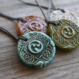 May include: Four circular ceramic pendants on dark cords, each featuring a central triskelion symbol surrounded by an intricate embossed knotwork pattern. The pendants are glazed in distinct colors including teal, olive green, brown, and white.