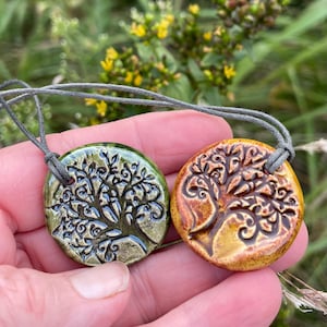 May include: Two circular ceramic pendants on grey cord necklaces. One pendant features a glossy olive green glaze with a dark embossed tree design, while the other has a warm amber orange glaze with a matching dark embossed tree motif.