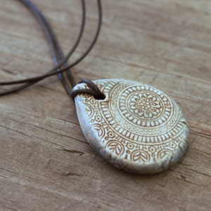 May include: Cream-colored, teardrop-shaped pendant with intricate floral and geometric patterns. The pendant is suspended from a dark brown cord. The design features detailed relief work, creating a textured surface. A unique, handcrafted necklace.