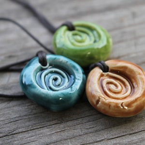 May include: Three ceramic pendant necklaces in shades of green, teal, and brown. Each pendant features a spiral design and is strung on a dark cord. The pendants are displayed on a weathered wooden surface.
