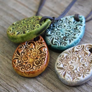 May include: Four ceramic pendant necklaces in shades of green, teal, brown, and off-white. Each pendant has a unique floral design and is strung on a dark brown cord. The pendants are displayed on a weathered wooden surface.