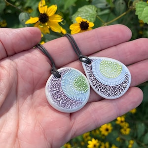 May include: Two ceramic pendants on dark gray cords. One pendant is circular and the other is teardrop-shaped. Both feature a white base with intricate stamped patterns in shades of purple, light blue, and lime green, showcasing a central floral motif.