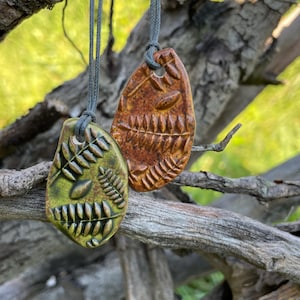 May include: Two teardrop-shaped ceramic pendants hanging from grey cords. One pendant is olive green and the other is reddish-brown. Both feature embossed fern leaf patterns and small oval shapes on their surfaces, with a hole at the top for the cord.