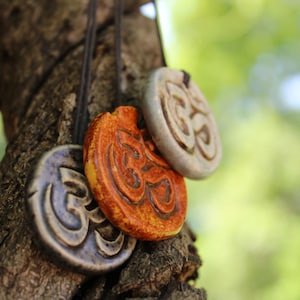 May include: Close-up of four ceramic Om symbol pendants in shades of blue, orange, and gray, hanging from a tree branch. The pendants are round with a raised Om symbol design and are strung on black cords. The background is blurred green.