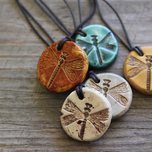 May include: A collection of handmade dragonfly pendant necklaces. Each pendant is a different color, including orange, teal, and cream, with a detailed dragonfly design. The pendants are strung on black cords, perfect for everyday wear.